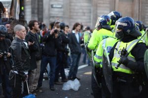 Image of two opposing lines; on the left, a group of protestors, on the right a group of police in riot gear.